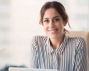 Eine Frau mit langen, braunen Haaren sitzt an einem Schreibtisch und lächelt. Sie trägt ein gestreiftes Hemd und hat einen Laptop vor sich. Im Hintergrund sind unscharfe Pflanzen und Fenster zu sehen.