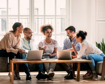 Fünf Personen sitzen um einen Tisch mit Laptops, lachen und diskutieren. Sie scheinen an einem kreativen Projekt oder einer Besprechung zu arbeiten. Der Raum hat große Fenster und eine helle, einladende Atmosphäre.