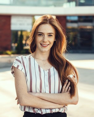 Junge Frau mit langen, lockigen Haaren und gestreiftem Oberteil, die lächelnd mit verschränkten Armen in einem urbanen Umfeld steht.