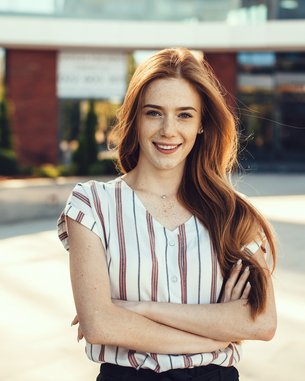 Junge Frau mit langen, lockigen Haaren und gestreiftem Oberteil, die lächelnd mit verschränkten Armen in einem urbanen Umfeld steht.