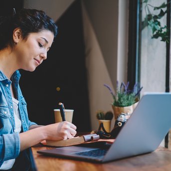 A woman sits at a table, writing in a notebook and working on a laptop while enjoying a cup of coffee.