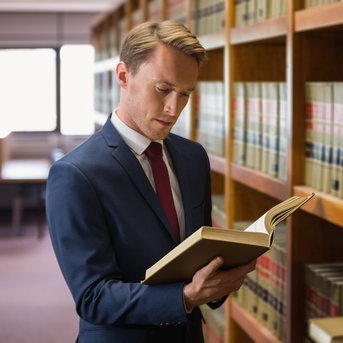 Ein Mann in einem Anzug steht in einer Bibliothek und liest ein Buch, während er an einem Regal mit juristischen Fachbüchern lehnt.