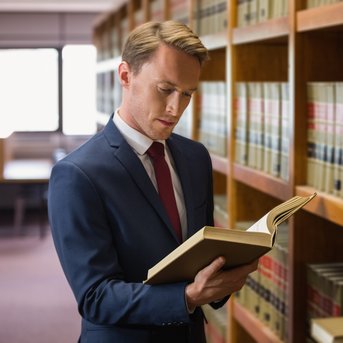 Ein Mann in einem Anzug steht in einer Bibliothek und liest ein Buch, während er an einem Regal mit juristischen Fachbüchern lehnt.