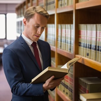 Ein Mann in einem Anzug steht in einer Bibliothek und liest ein Buch, während er an einem Regal mit juristischen Fachbüchern lehnt.