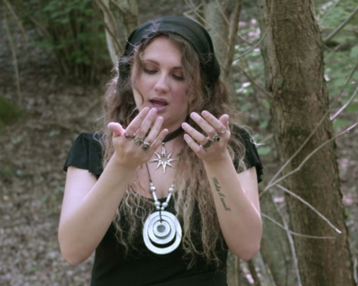 A woman with long, curly hair and jewelry stands in the forest, looking at her hands. She is wearing a black top and a hat.