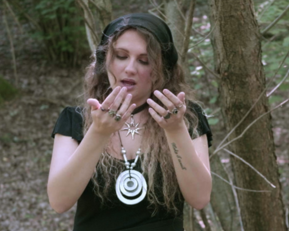 A woman with long, curly hair and jewelry stands in the forest, looking at her hands. She is wearing a black top and a hat.