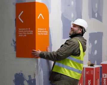 A construction worker wearing a white helmet and a reflective vest is installing an orange sign with information about seminars and media studios.