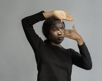 Eine Person in einem schwarzen Oberteil hält eine Hand über ihr Gesicht und zeigt mit der anderen Hand nach vorne.