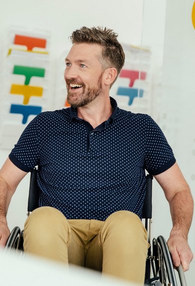 Ein Mann mit einem Bart sitzt in einem Rollstuhl und lächelt. Er trägt ein gepunktetes Hemd und beige Hosen. Im Hintergrund sind farbige Muster oder Farbmuster sichtbar.