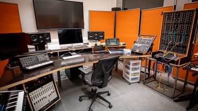 Ein modernes Musikstudio mit Synthesizern, Lautsprechern und einem Computer. Der Raum hat orangefarbene Wände und eine große Bildschirmfläche. Ein Drehstuhl steht an einem geschwungenen Tisch.