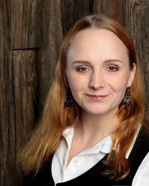 Eine Frau mit langen, roten Haaren und Ohrringen lächelt in die Kamera, vor einer rustikalen Holzoberfläche.