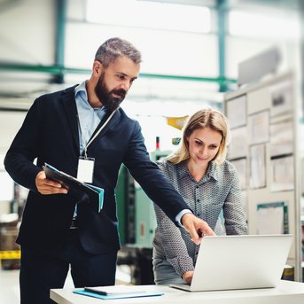A man and a woman are standing at a table with a laptop. The man is pointing at the screen while the woman is watching attentively.