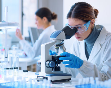 Eine Wissenschaftlerin betrachtet Proben durch ein Mikroskop, während eine weitere Person im Hintergrund an einem Tisch arbeitet.