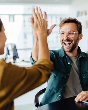 Ein Mann gibt einer Frau ein High-Five, während eine weitere Frau am Tisch lächelt. Ein Laptop steht auf dem Tisch.