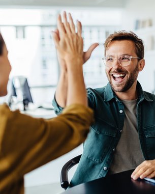 Ein Mann gibt einer Frau ein High-Five, während eine weitere Frau am Tisch lächelt. Ein Laptop steht auf dem Tisch.