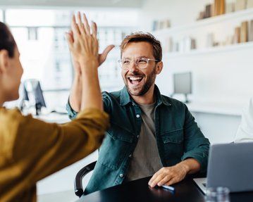 Ein Mann gibt einer Frau ein High-Five, während eine weitere Frau am Tisch lächelt. Ein Laptop steht auf dem Tisch.