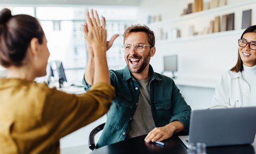 Ein Mann gibt einer Frau ein High-Five, während eine weitere Frau am Tisch lächelt. Ein Laptop steht auf dem Tisch.