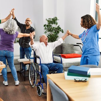 Eine Gruppe von fünf Personen, darunter ein älterer Mann im Rollstuhl, tanzt und hebt die Hände in einem hellen Raum.