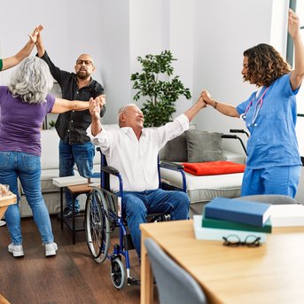 A group of five people, including an older man in a wheelchair, is dancing and raising their hands in a bright room.