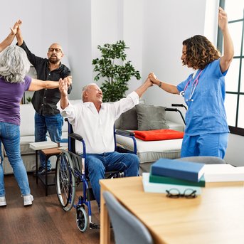 Eine Gruppe von fünf Personen, darunter ein älterer Mann im Rollstuhl, tanzt und hebt die Hände in einem hellen Raum.