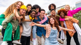 Eine Gruppe von zwölf jungen Menschen posiert fröhlich im Freien, einige tragen bunte Kleidung und lächeln in die Kamera.