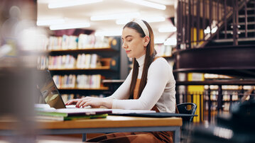 Eine junge Frau sitzt an einem Tisch in einer Bibliothek und arbeitet konzentriert an einem Laptop. Bücherregale sind im Hintergrund sichtbar.