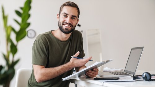 Ein Mann mit Bart sitzt an einem Schreibtisch, hält einen Stift und einen Notizblock, während er in einen Laptop schaut.