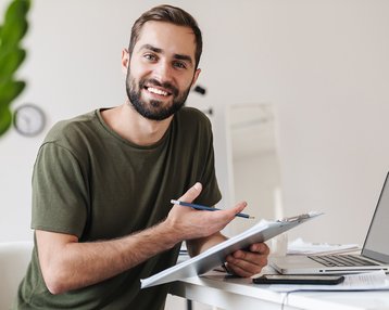 Ein Mann mit Bart sitzt an einem Schreibtisch, hält einen Stift und einen Notizblock, während er in einen Laptop schaut.