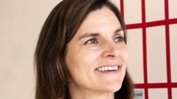 Eine Frau mit langen, braunen Haaren lächelt und schaut nach rechts. Sie trägt ein schwarzes Oberteil mit weißen Details. Im Hintergrund sind rote Gitterelemente sichtbar.