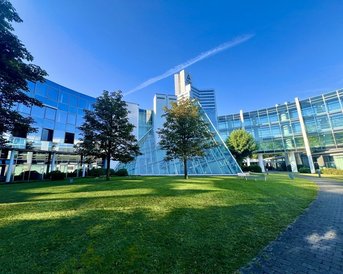Moderne Architektur mit einer gläsernen Pyramide und umliegenden Bürogebäuden, umgeben von Rasenflächen und Bäumen. Der Himmel ist klar und blau.