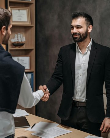 Zwei Männer schütteln sich die Hände in einem Büro. Der Mann im Anzug lächelt, während der andere, leger gekleidet, ihm gegenübersteht. Auf dem Tisch liegen Dokumente und ein Stuhl ist im Hintergrund sichtbar.