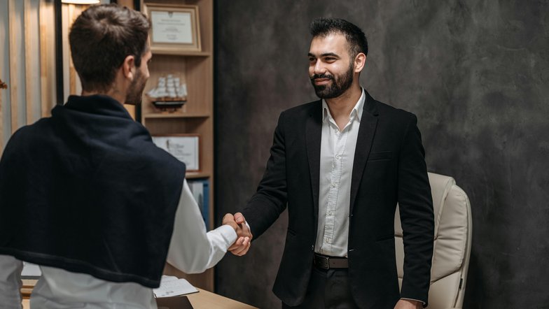 Zwei Männer schütteln sich die Hände in einem Büro. Der Mann im Anzug lächelt, während der andere, leger gekleidet, ihm gegenübersteht. Auf dem Tisch liegen Dokumente und ein Stuhl ist im Hintergrund sichtbar.