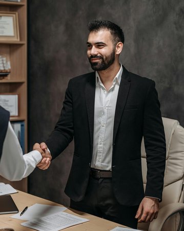 Zwei Männer schütteln sich die Hände in einem Büro. Der Mann im Anzug lächelt, während der andere, leger gekleidet, ihm gegenübersteht. Auf dem Tisch liegen Dokumente und ein Stuhl ist im Hintergrund sichtbar.
