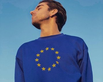 Junger Mann mit kurzem Haar trägt einen blauen Pullover mit dem EU-Sternenkranz, schaut nach oben vor einem klaren Himmel.