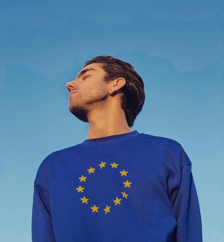 Junger Mann mit kurzem Haar trägt einen blauen Pullover mit dem EU-Sternenkranz, schaut nach oben vor einem klaren Himmel.
