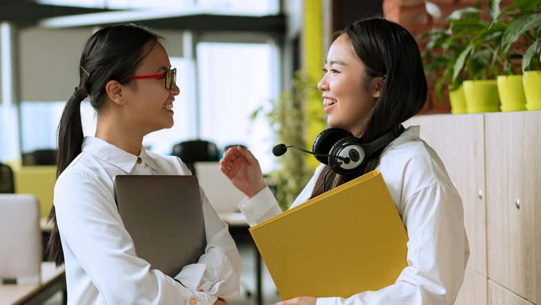 Zwei Frauen unterhalten sich lächelnd in einem modernen Büro. Eine hält einen Laptop, die andere hat einen Kopfhörer und eine gelbe Mappe in der Hand. Pflanzen und Büromöbel sind im Hintergrund sichtbar.