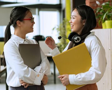 Kategorie Kommunikation SRH University Zwei Frauen unterhalten sich lächelnd in einem modernen Büro. Eine hält einen Laptop, die andere hat einen Kopfhörer und eine gelbe Mappe in der Hand. Pflanzen und Büromöbel sind im Hintergrund sichtbar.