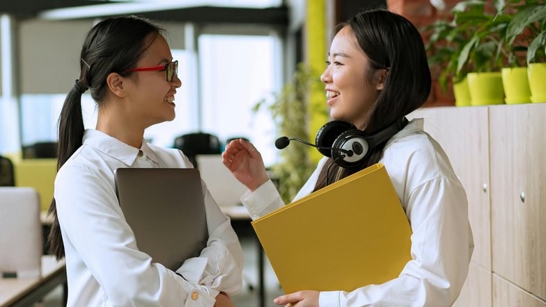 Zwei Frauen unterhalten sich lächelnd in einem modernen Büro. Eine hält einen Laptop, die andere hat einen Kopfhörer und eine gelbe Mappe in der Hand. Pflanzen und Büromöbel sind im Hintergrund sichtbar.