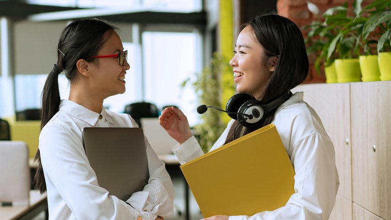 Zwei Frauen unterhalten sich lächelnd in einem modernen Büro. Eine hält einen Laptop, die andere hat einen Kopfhörer und eine gelbe Mappe in der Hand. Pflanzen und Büromöbel sind im Hintergrund sichtbar.