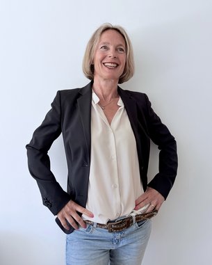 A woman with shoulder-length blonde hair is wearing a black blazer and a white blouse, standing with her hands on her hips in front of a white wall.
