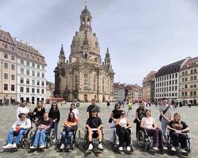 A group of people in wheelchairs gathered in a square in front of a historic church, surrounded by buildings in the background.