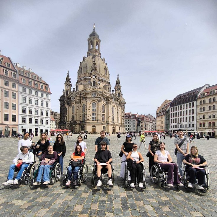 A group of people in wheelchairs gathered in a square in front of a historic church, surrounded by buildings in the background.