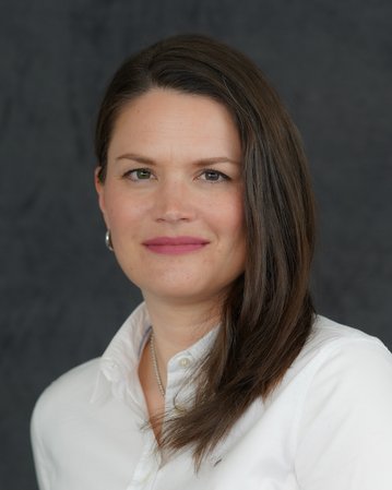 Eine Frau mit langen, braunen Haaren trägt ein weißes Hemd und lächelt leicht vor einem neutralen, grauen Hintergrund.