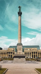 A tall column monument with a statue on top stands in the foreground, while in the background, a historic building with columns and a large roof is visible. People are moving around the area near the monument.
