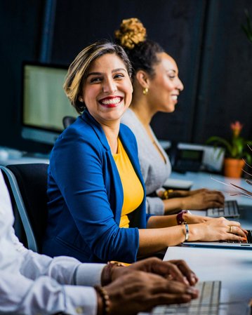 Drei Personen sitzen an einem Tisch in einem modernen Büro. Eine Frau lächelt freundlich in die Kamera, während die anderen konzentriert an ihren Laptops arbeiten. Pflanzen sind auf dem Tisch platziert.
