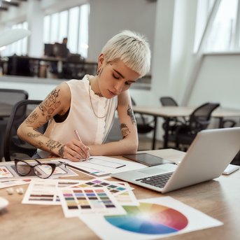 Eine tätowierte Frau mit kurzem, blonden Haar arbeitet an einem Designprojekt, umgeben von Farbmustern und einem Laptop auf einem Tisch.
