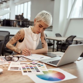 A tattooed woman with short blonde hair is working on a design project, surrounded by color swatches and a laptop on a table.