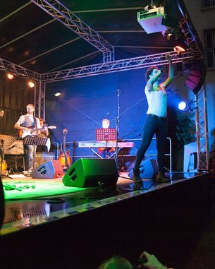 Ein Musiker tritt auf einer Bühne auf, umgeben von Instrumenten und Lichtern. Im Hintergrund sind weitere Bandmitglieder sichtbar. Das Publikum beobachtet die Darbietung aufmerksam.