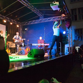 Ein Musiker tritt auf einer Bühne auf, umgeben von Instrumenten und Lichtern. Im Hintergrund sind weitere Bandmitglieder sichtbar. Das Publikum beobachtet die Darbietung aufmerksam.