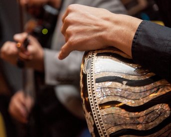 Eine Hand spielt auf einer Trommel mit dekorativer Oberfläche, während im Hintergrund ein Musiker mit einer Violine zu sehen ist.